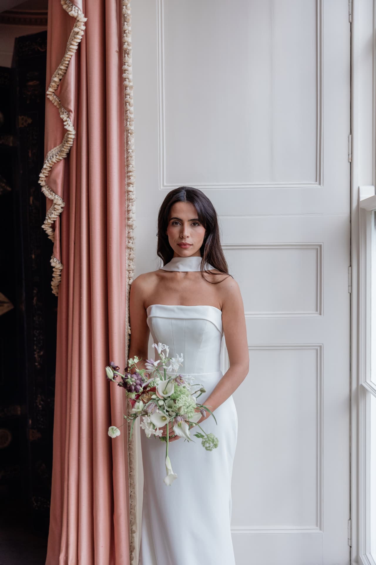 Bride in a sculpted white gown holding a spring bouquet beside a silk curtain.