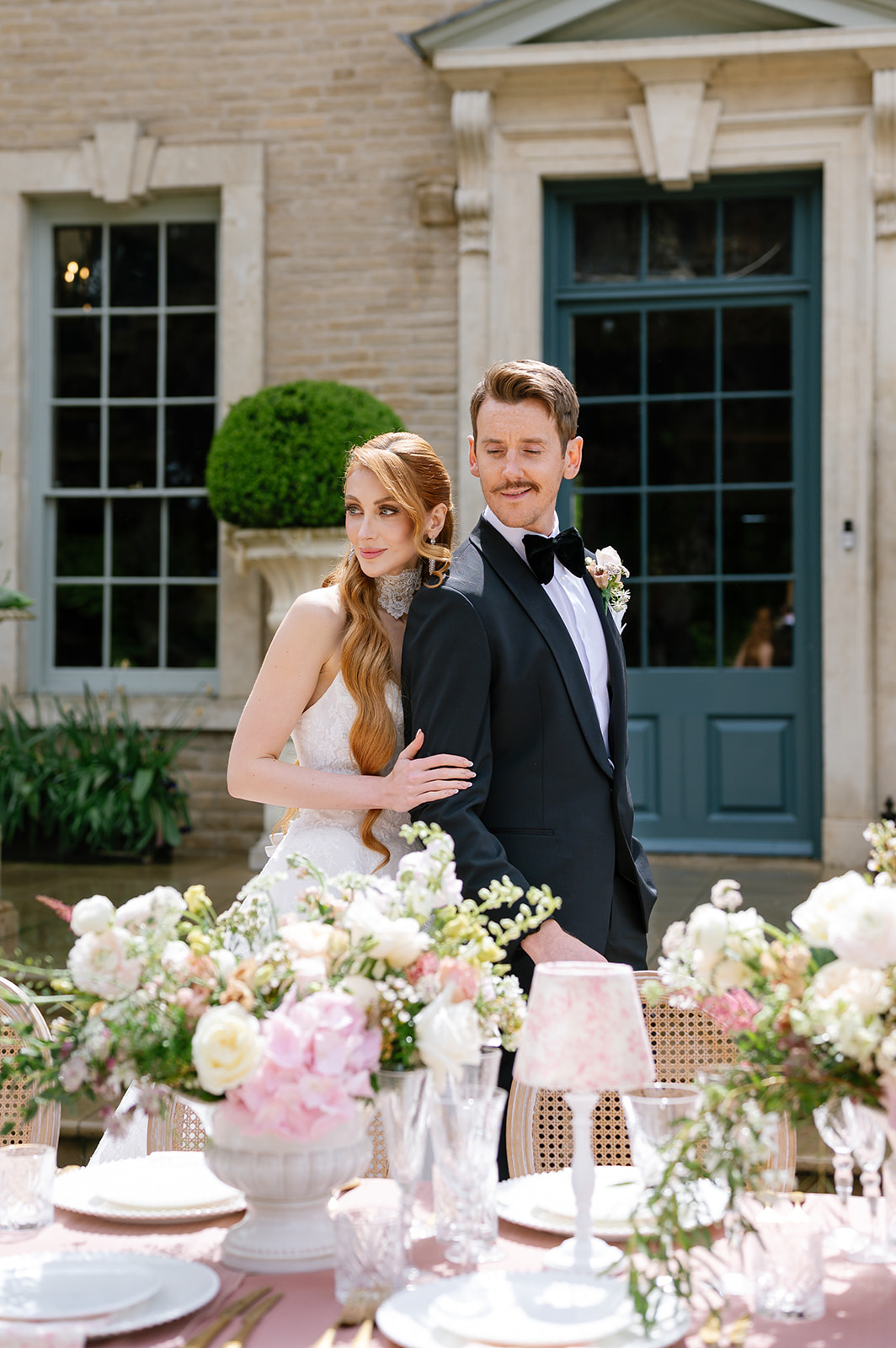 Bride and groom standing behind a dressed outdoor table at a stone house.