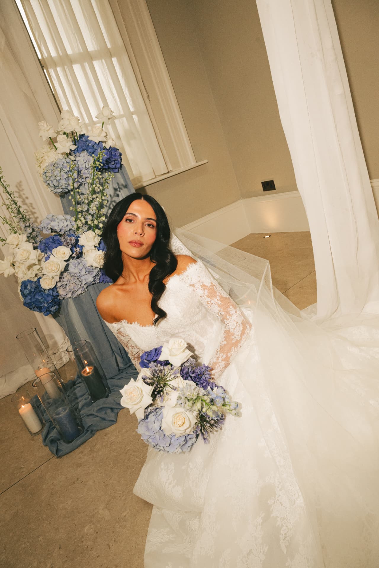 Bride in a lace-sleeved gown seated beside a blue hydrangea installation.