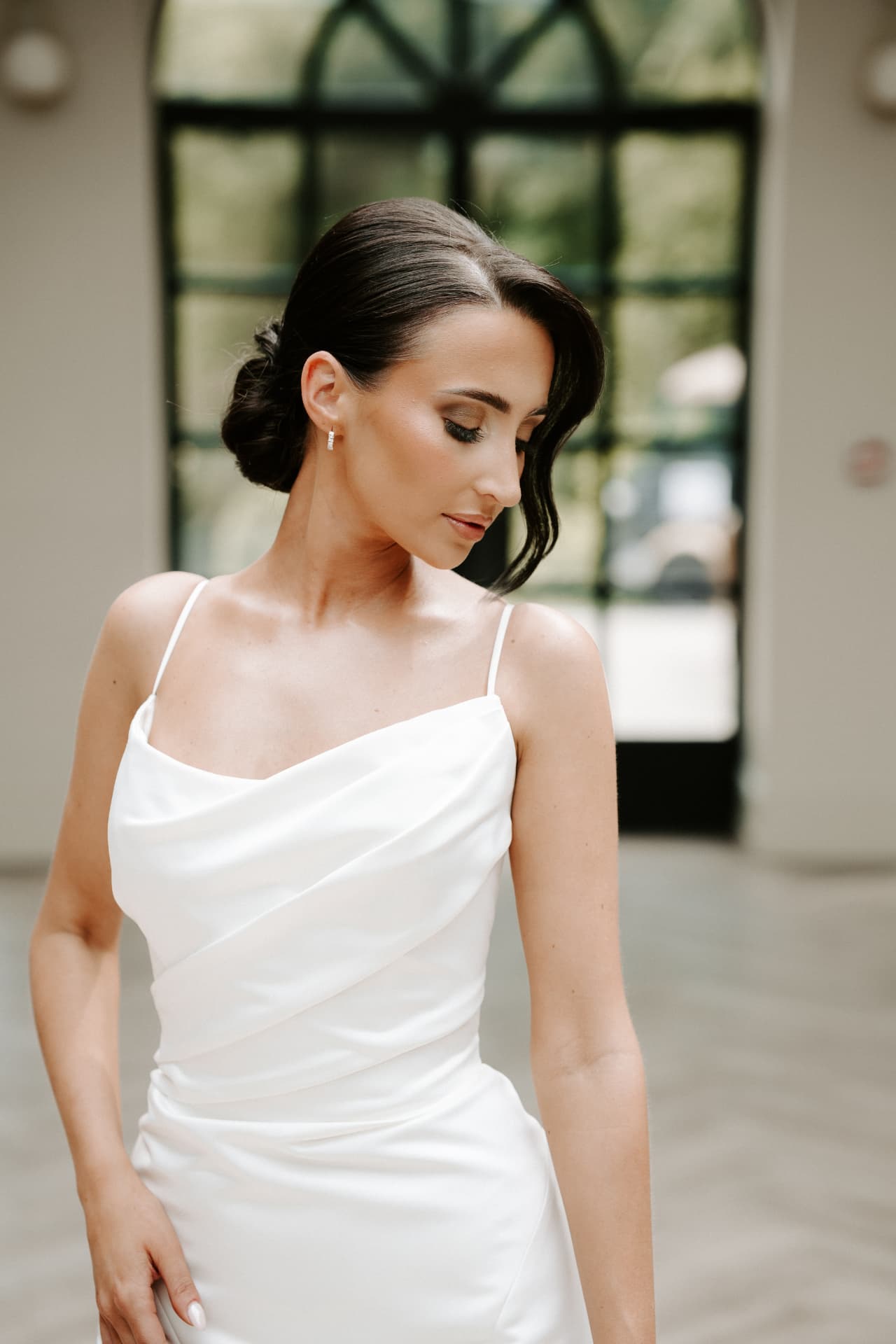 Bride in a cowl-neck gown with a smooth low bun, looking down.