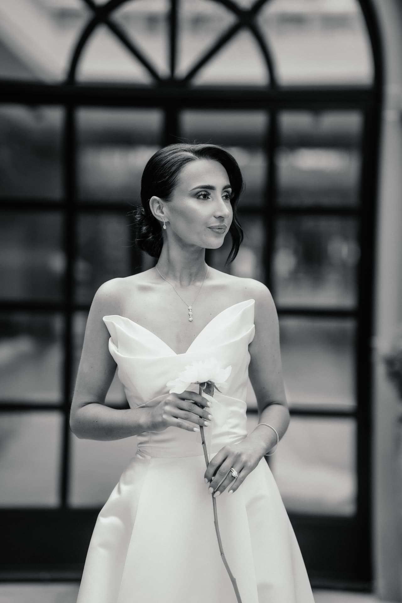 Black and white portrait of a bride holding a peony in front of a tall glazed door.